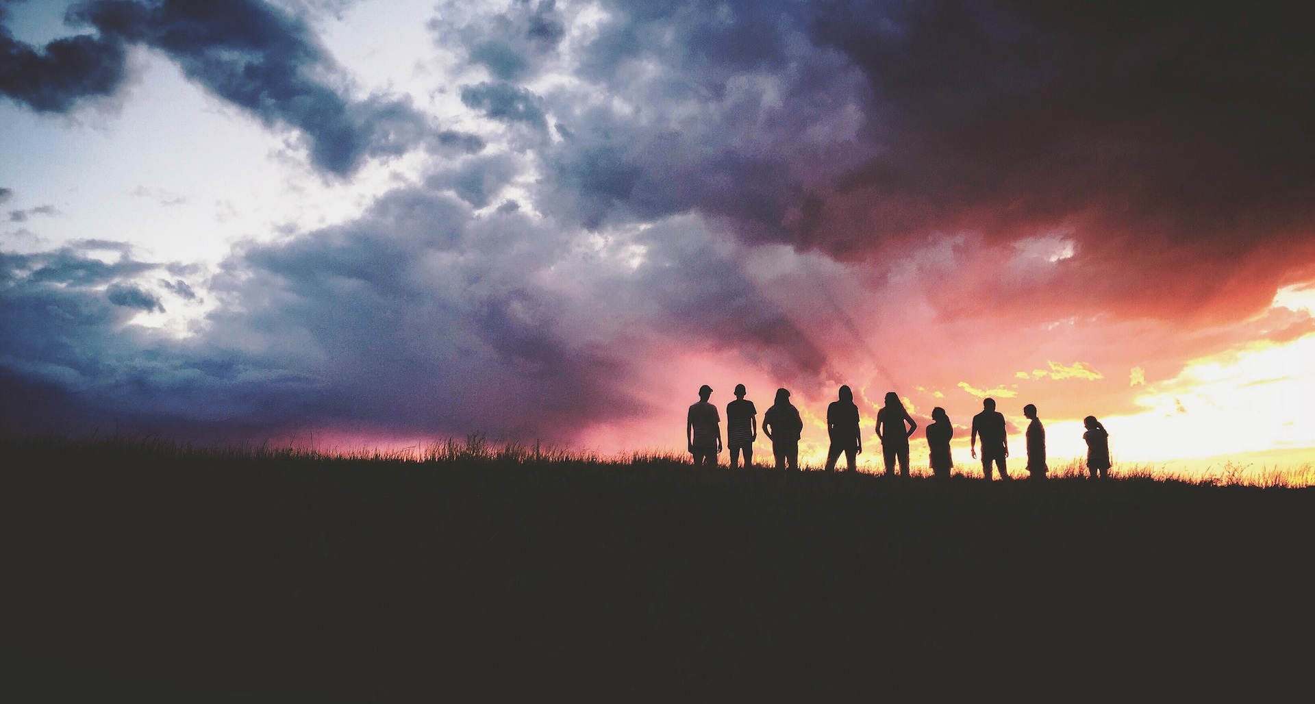 A group of people silhouetted on a grassy hilltop under a stormy sunset sky
